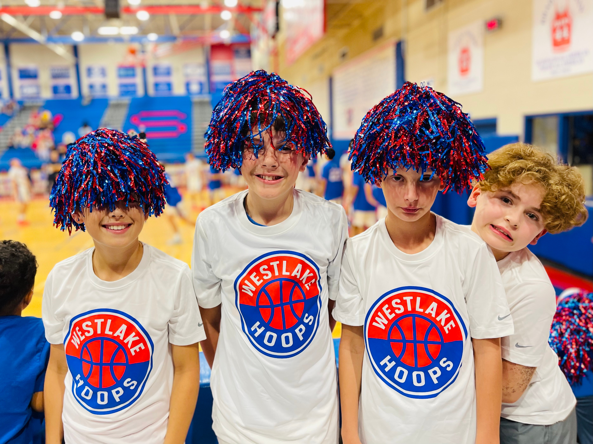 Four Westlake Hoops players wearing pom-pom wigs courtside at a Chaps game.